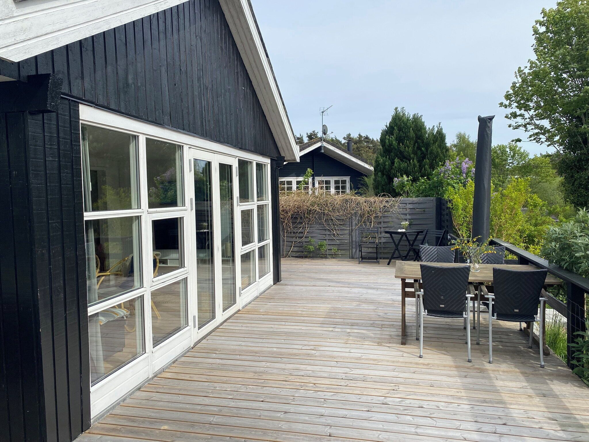 En træterrasse med et bord og stole, en stor skydedør samt en bygning med mørke vægge og et skråt tag.