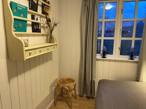 Ein Zimmer mit Bücherregal, einem Korbstuhl, einem Fenster mit Vorhängen und einem Bett.