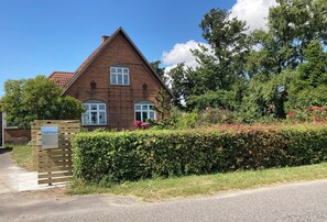 Une maison en briques avec un toit triangulaire, une porte en bois et une haie devant.