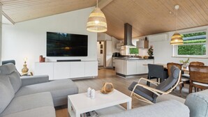 A modern living room with a sofa, coffee table, and a TV. There is a dining area with chairs and a kitchen in the background. - Lovely holiday home Spa/sauna, carcharger, river (Fårvang)