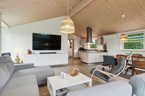 Una sala de estar moderna con un sofá, una mesa de centro y un televisor. Se observa un comedor con sillas y una cocina al fondo.