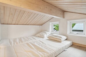 Un dormitorio con techo de madera, una cama con ropa de cama blanca y dos ventanas que dan a zonas verdes.