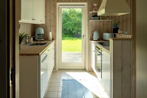 A kitchen with wooden cabinets, a coffee maker, a toaster, and a view of a green lawn through an open door. - Charming house in the heart of WestZealand (Eskebjerg)