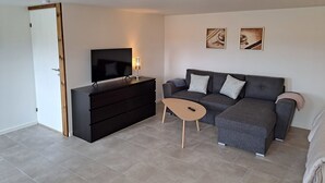 A modern living room with a flat-screen TV, a dark wooden dresser, a grey sofa with two cushions, and a small wooden side table.