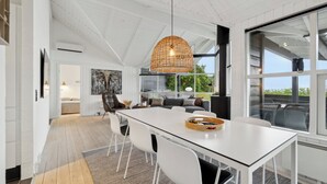Een modern interieur met een glazen eettafel, witte stoelen, een geweven hanglamp en uitzicht op de omgeving door grote ramen.