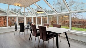 A modern dining area with a glass ceiling, wooden beams, and a view of trees and a brick wall.