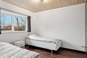 A bedroom with a single bed, a small bedside table, a window with curtains, and a view of trees and buildings outside.