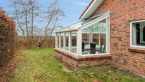 A house with a glass conservatory attached to the side, a brick exterior, and a grassy yard with trees.