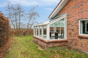 A house with a glass conservatory attached to the side, a brick exterior, and a grassy yard with trees.