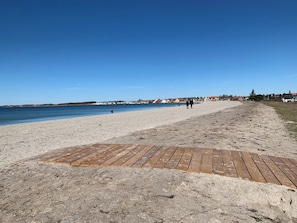 Una playa con una pasarela de madera que lleva al agua, cielo despejado y casas al fondo.