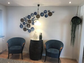 Une pièce avec deux fauteuils bleus, une table ronde ornée d’un objet décoratif, un mur recouvert d’assiettes bleues et brunes, et une plante suspendue.