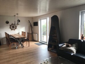 Een woonkamer met een houten tafel, stoelen, een zwarte leren bank, een open haard en een groot raam.