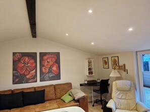 A living room with a brown sofa, a white armchair, a desk with a chair, and two paintings on the wall.