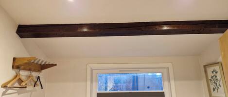 A room with a wooden ceiling beam, a window with a view of trees, a door with a vent, and a wall-mounted light fixture.