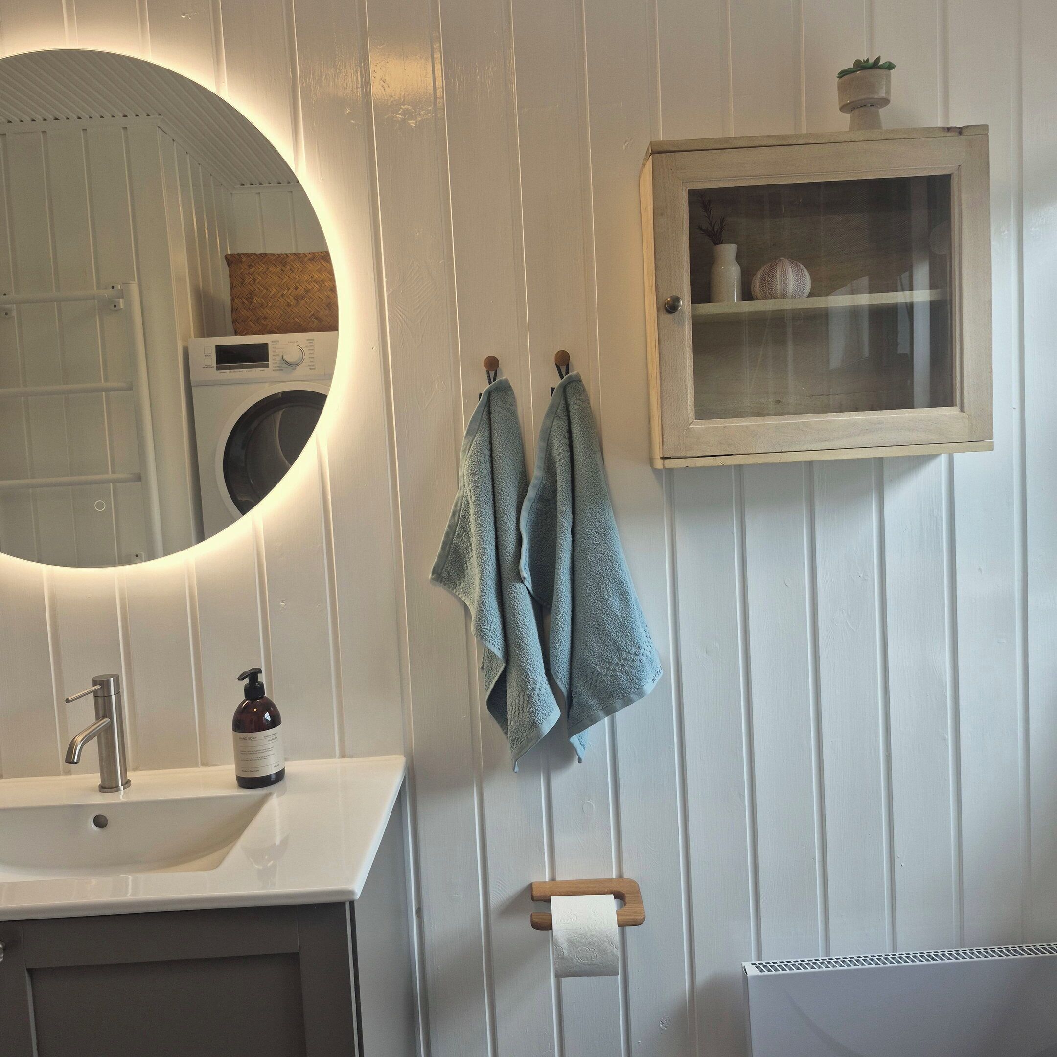 Une salle de bain avec un miroir rond, un lavabo avec un robinet, un flacon de savon liquide, un porte-serviettes avec des serviettes, un porte-rouleau de papier toilette et un meuble avec une porte en verre.