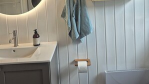 Ein Badezimmer mit rundem Spiegel, Waschbecken mit Wasserhahn, Handseifenflasche, Handtuchhalter mit Handtüchern, Toilettenpapierhalter und einem Schrank mit Glastür.
