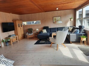 A modern living room with a wooden ceiling, a large flat-screen TV, a dark sofa, a light armchair, and a wooden coffee table.