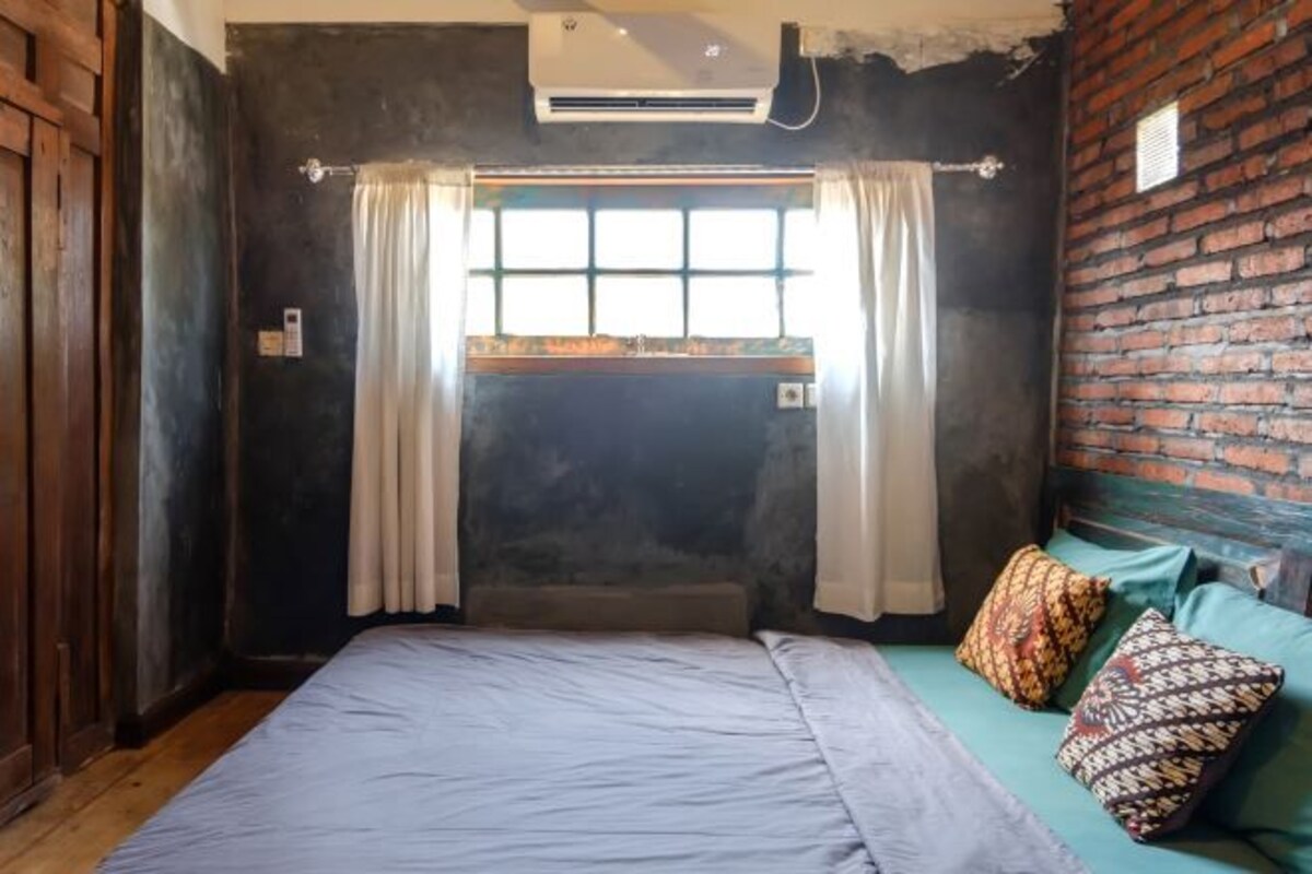A room with a bed, a window with curtains, a brick wall, and a wooden door.