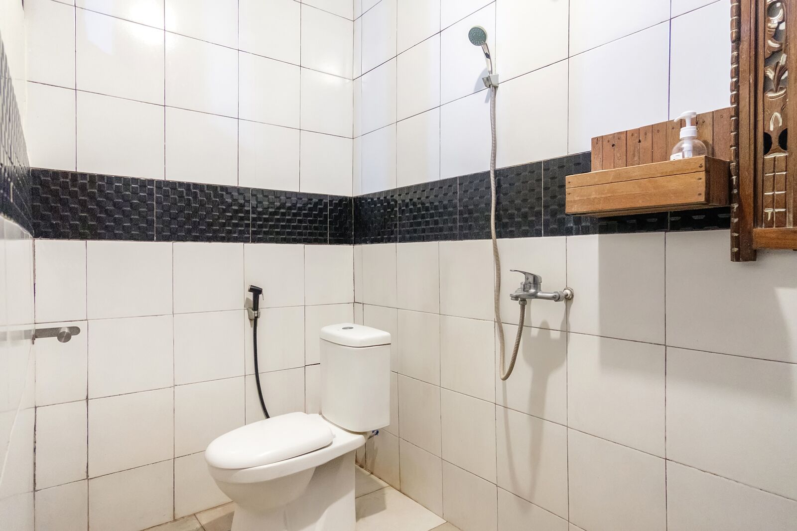 A bathroom with white tiled walls, a black and white patterned accent wall, a white toilet, a shower with a handheld showerhead, and a wooden shelf with a bottle.