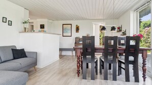 Een moderne woonkamer met een grijze bank, een houten eettafel en stoelen, een witte muur met ingelijste foto’s en uitzicht op groen buiten.