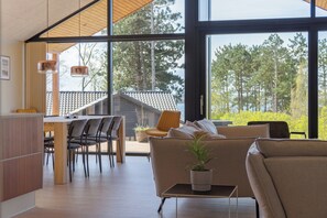 A modern interior with a dining area, sofa, and large windows offering a view of trees and a building. - Impressive holiday cottage by the Helsnæs Bay (Millinge)