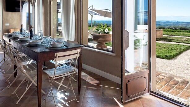 Un comedor con una mesa larga dispuesta para seis personas, amplias ventanas en arco y vistas al campo.