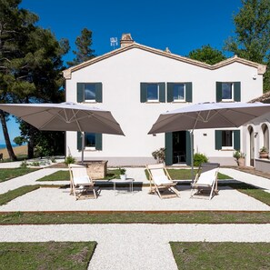 Un bâtiment blanc à deux étages, doté d’une façade symétrique, avec une entrée centrale flanquée de deux grands parasols et de trois fauteuils de jardin.