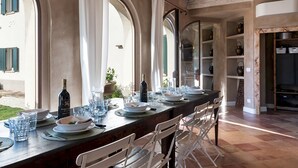Una larga mesa de comedor dispuesta con sillas, cortinas blancas y vistas al exterior.