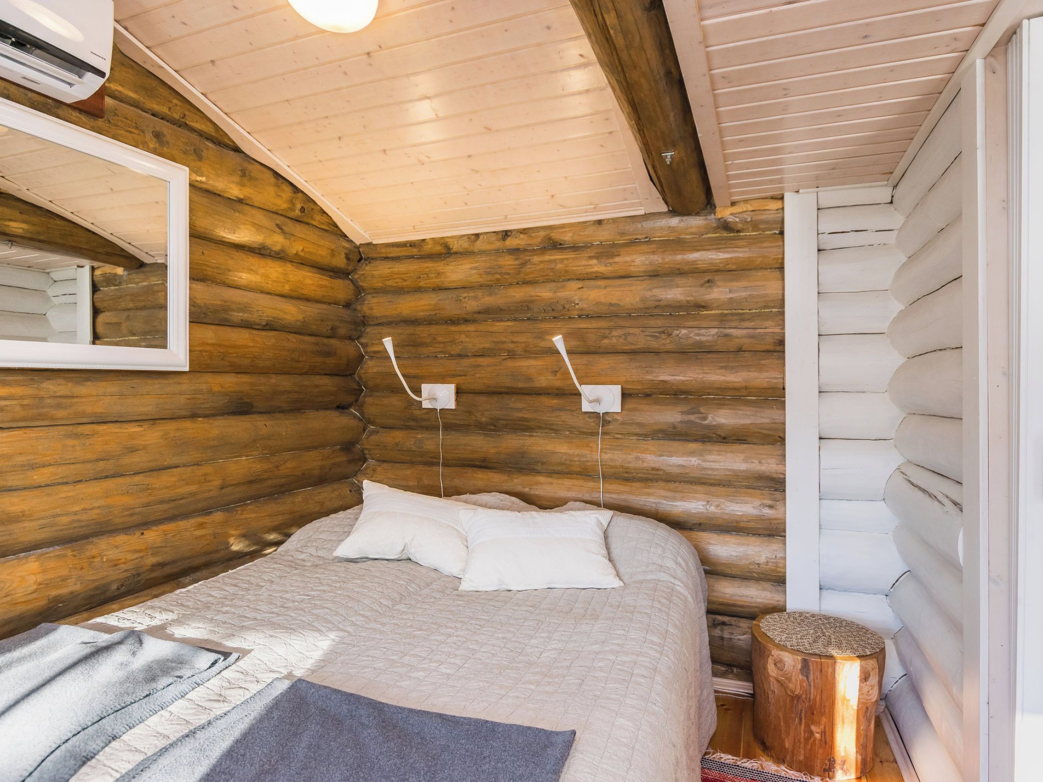 Een slaapkamer in een blokhut, met een bed, kussens, een nachtkastje, een spiegel en een wandlamp.