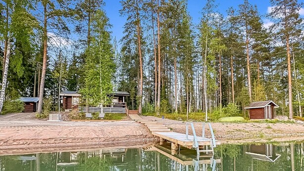 Eine Holzhütte mit einem Steg, der in einen stillen See hineinreicht, umgeben von hohen Kiefern.