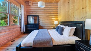 Une chambre à coucher située dans une cabane en rondins, avec un lit, une table de chevet, une lampe, une commode, un miroir et de grandes fenêtres.