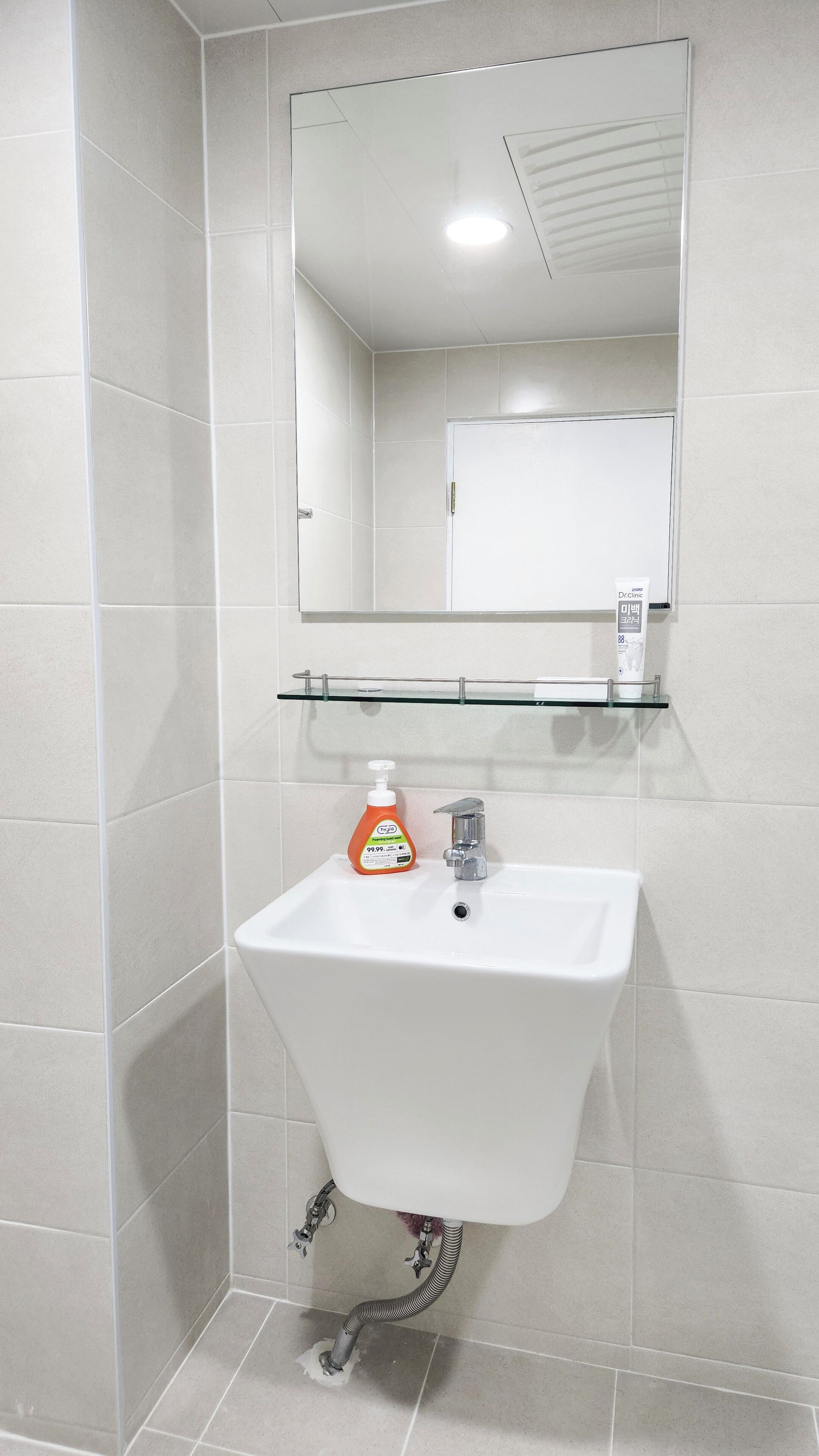 Een badkamer met een wit wastafelmeubel, een spiegel erboven en een glazen plankje met een flesje handdesinfecterend middel.