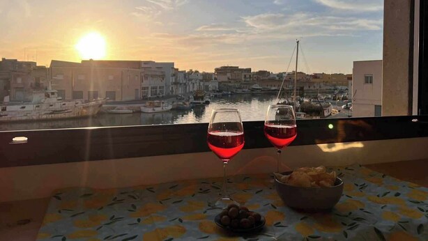 Un balcon avec une table dressée pour deux personnes, deux verres de vin rouge, un panier de pain et une vue sur une marina avec des bateaux et des bâtiments, le tout baigné par les couleurs du coucher de soleil.