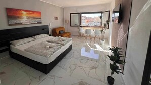 Une chambre moderne avec un grand lit, une vue sur la ville par la fenêtre et un coin repas avec des chaises.