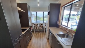A modern kitchen with dark cabinetry, a central island with a sink, and large windows offering a view of the outdoors. - Tidy Three Bedroom Home in Ruakaka. (Ruakākā)