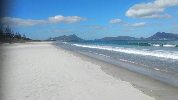 A beach with a clear sky, waves, and distant mountains. - Tidy Three Bedroom Home in Ruakaka. (Ruakākā)