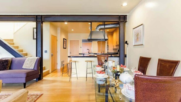 A modern living room with a sofa, dining table, and chairs, a kitchen in the background, and a staircase leading to another level. - Two  Bedroom, Two Newly Renovated Bathrooms, Deck (San Francisco)