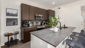 A modern kitchen with dark wood cabinets, stainless steel appliances, a central island with bar stools, and a potted plant. - The Macallister | Luxe 2BR Near TPC Scottsdale (Scottsdale)