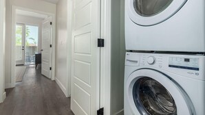 A laundry room with stacked front-loading washers and dryers, a hallway with white doors, and a view of the outside through a door. - The Macallister | Luxe 2BR Near TPC Scottsdale (Scottsdale)