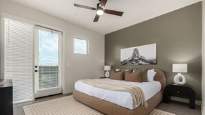 Une chambre à coucher comprenant un lit, deux tables de chevet avec des lampes, un ventilateur de plafond, une grande fenêtre avec des stores et une œuvre d’art accrochée au mur.