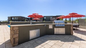 An outdoor bar area with a grill, stone counter, and red umbrellas. - The Macallister | Luxe 2BR Near TPC Scottsdale (Scottsdale)