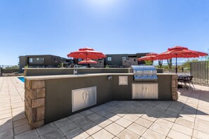 Un espace bar extérieur avec un barbecue, un comptoir en pierre et des parasols rouges.