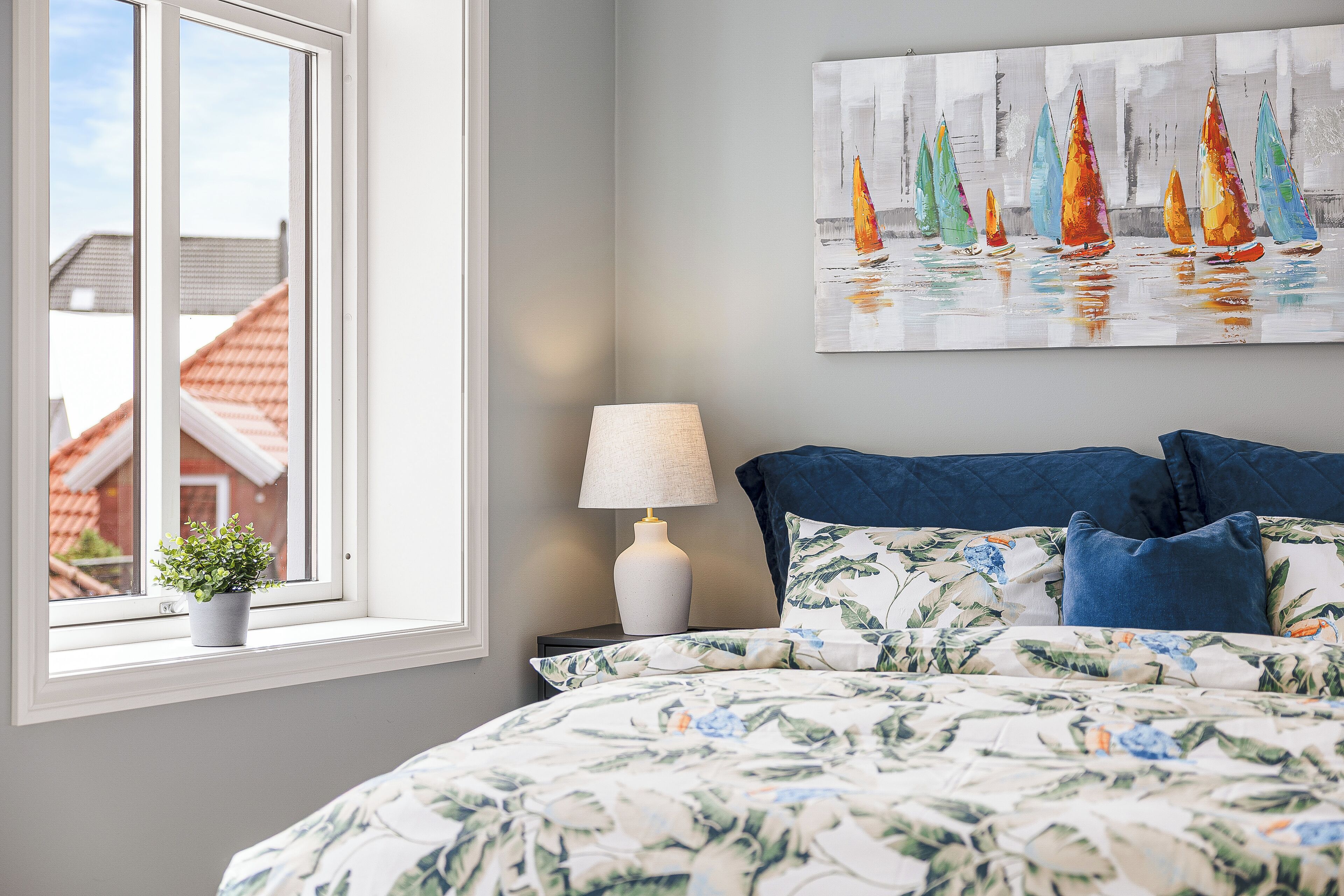 Een slaapkamer met een bed, een raam met uitzicht op huizen, een nachtkastje met een lamp en een schilderij aan de muur.