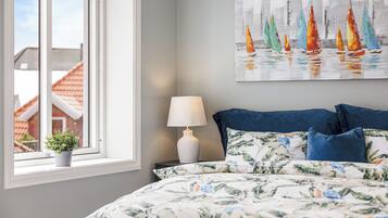 A bedroom with a bed, a window with a view of houses, a nightstand with a lamp, and a painting on the wall.