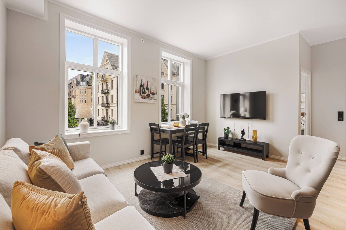 A modern living room with a sofa, armchair, round coffee table, dining table, and chairs. There is a TV mounted on the wall and a window with a view of buildings.