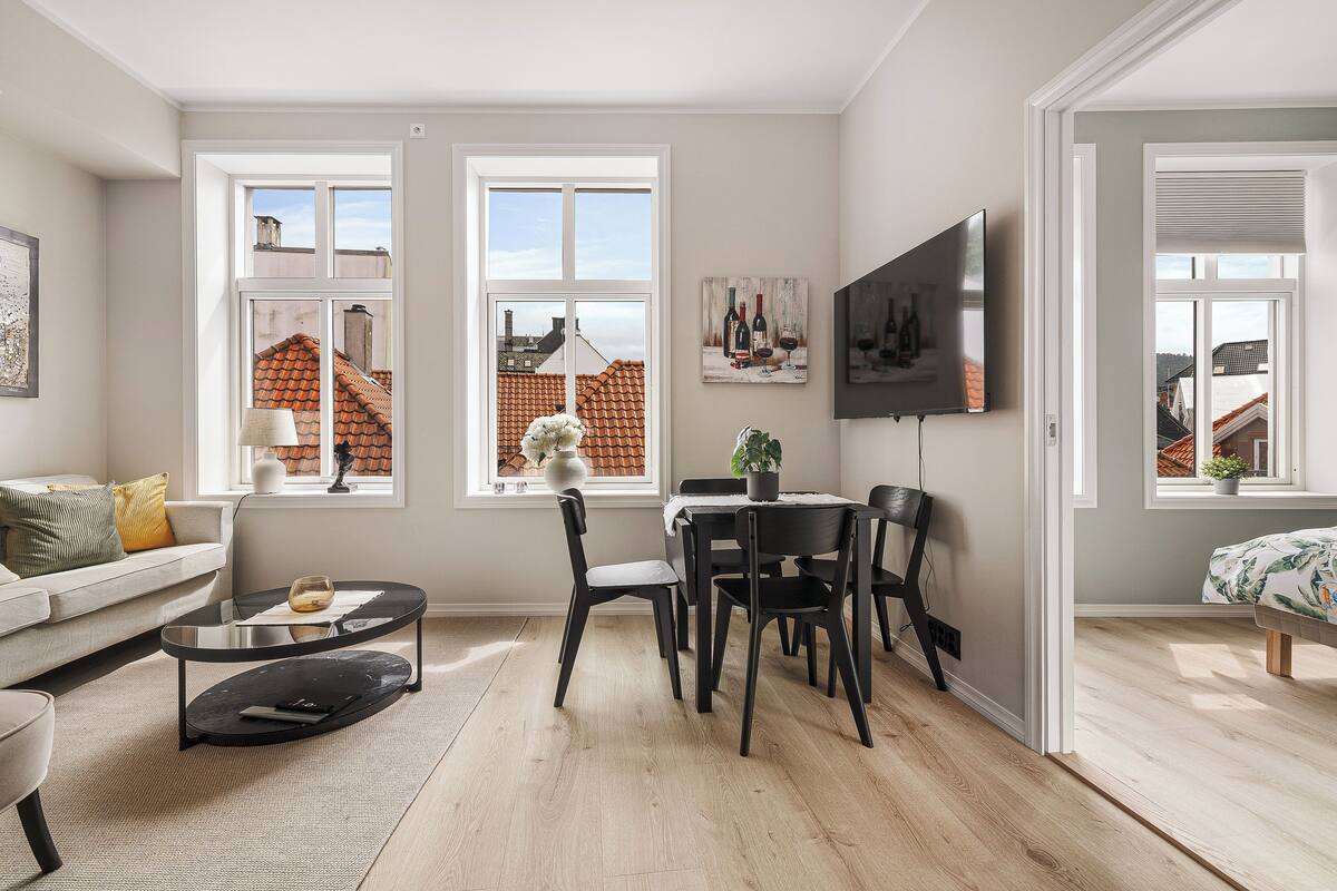 A modern living room with a sofa, a dining table with chairs, a coffee table, and a TV mounted on the wall. There are windows with views of buildings and a plant on the windowsill.