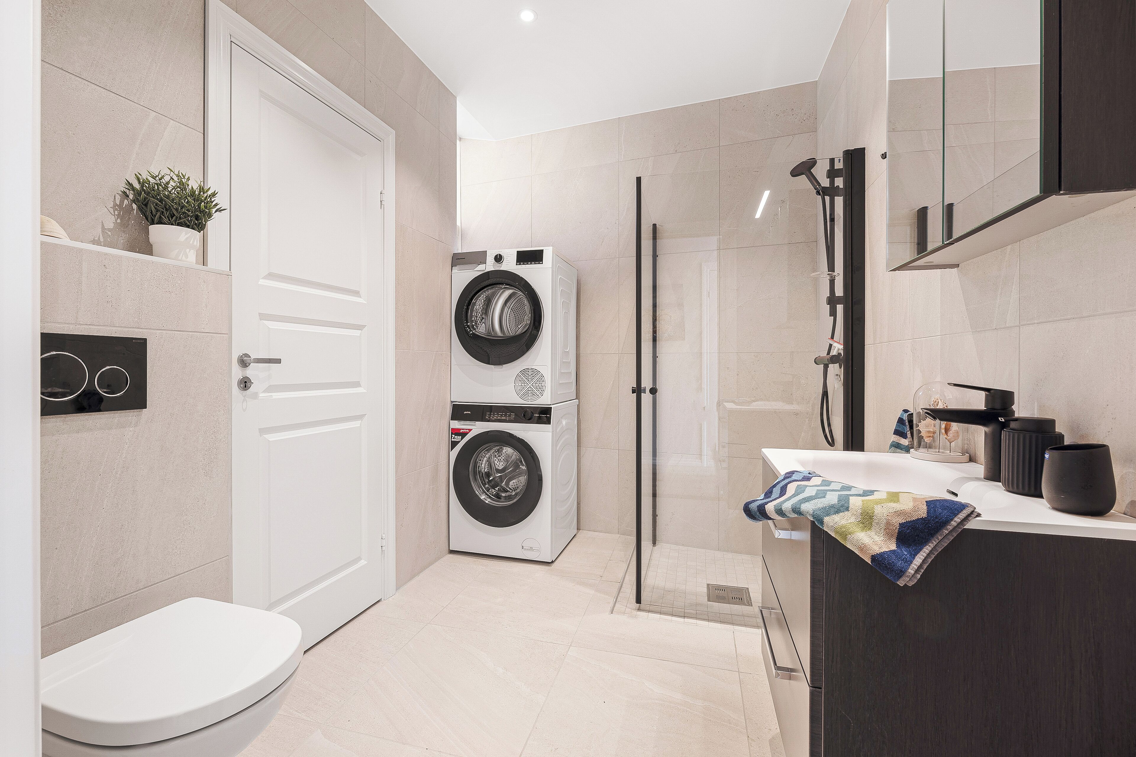 Ein modernes Badezimmer mit Toilette, einer Waschmaschine für zwei Personen, einer Dusche und einem Waschbecken mit Handtuch.