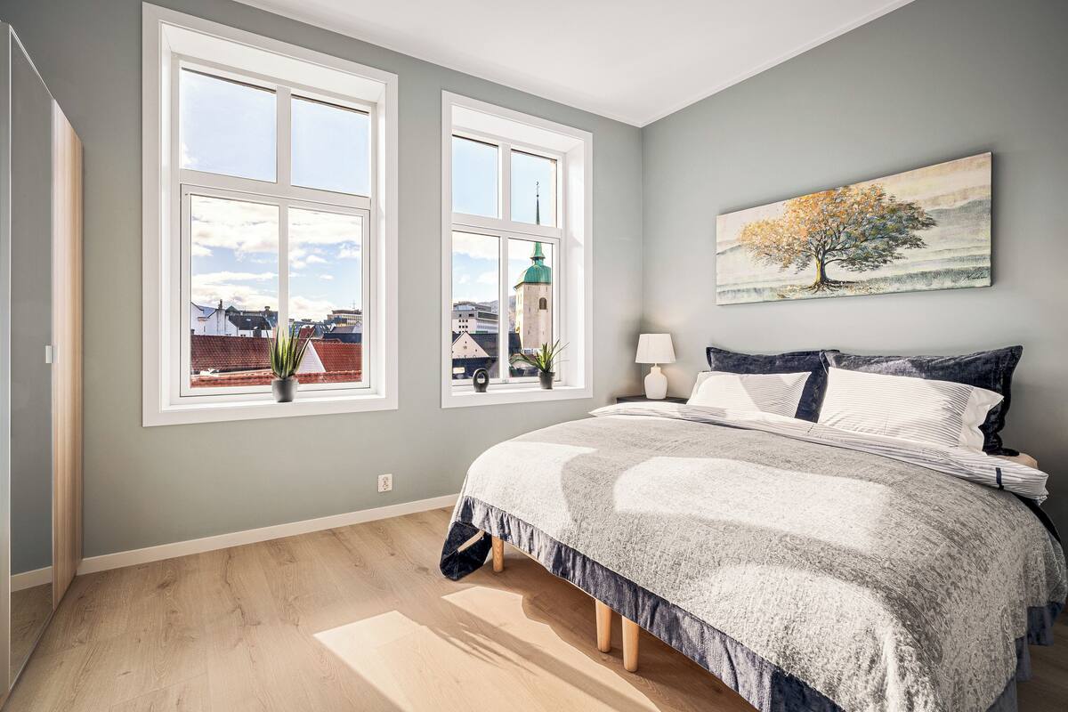 A bedroom with a large bed, two windows, a painting, and a view of buildings.