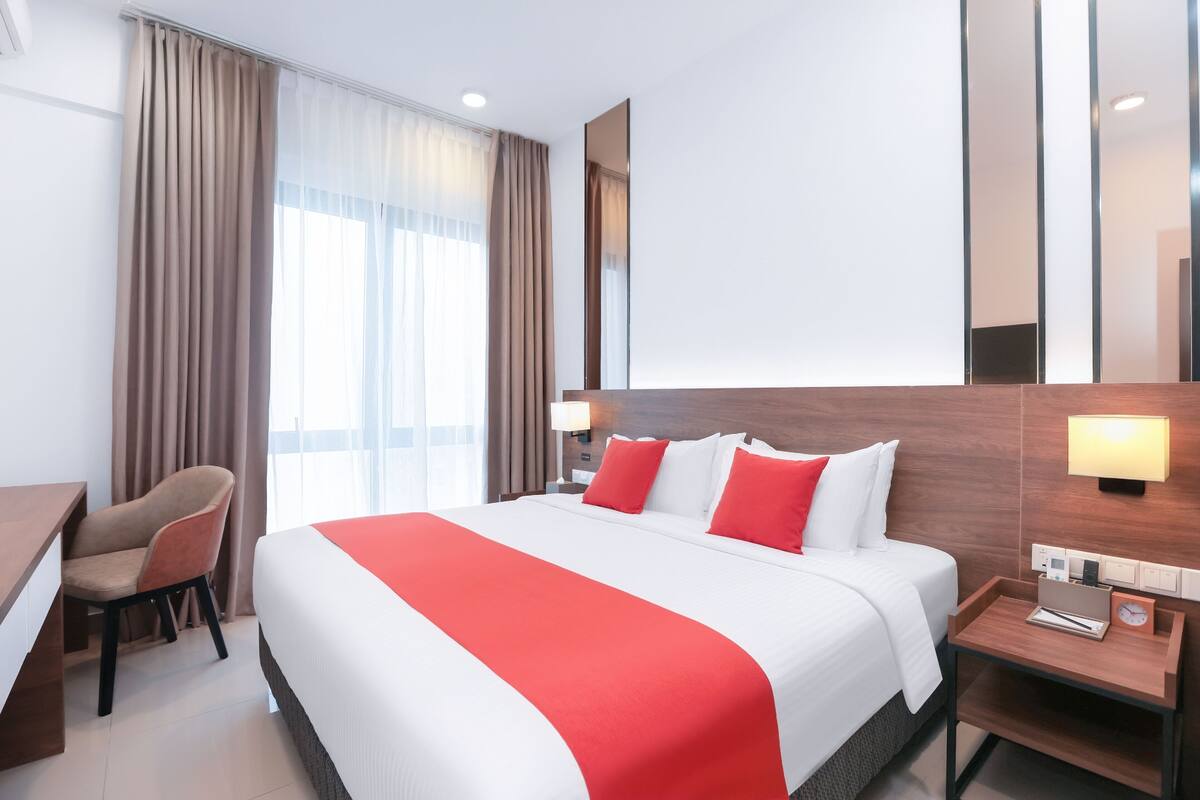 Habitación de hotel con una cama grande, un escritorio, una silla y una mesita de noche con lámpara y reloj.