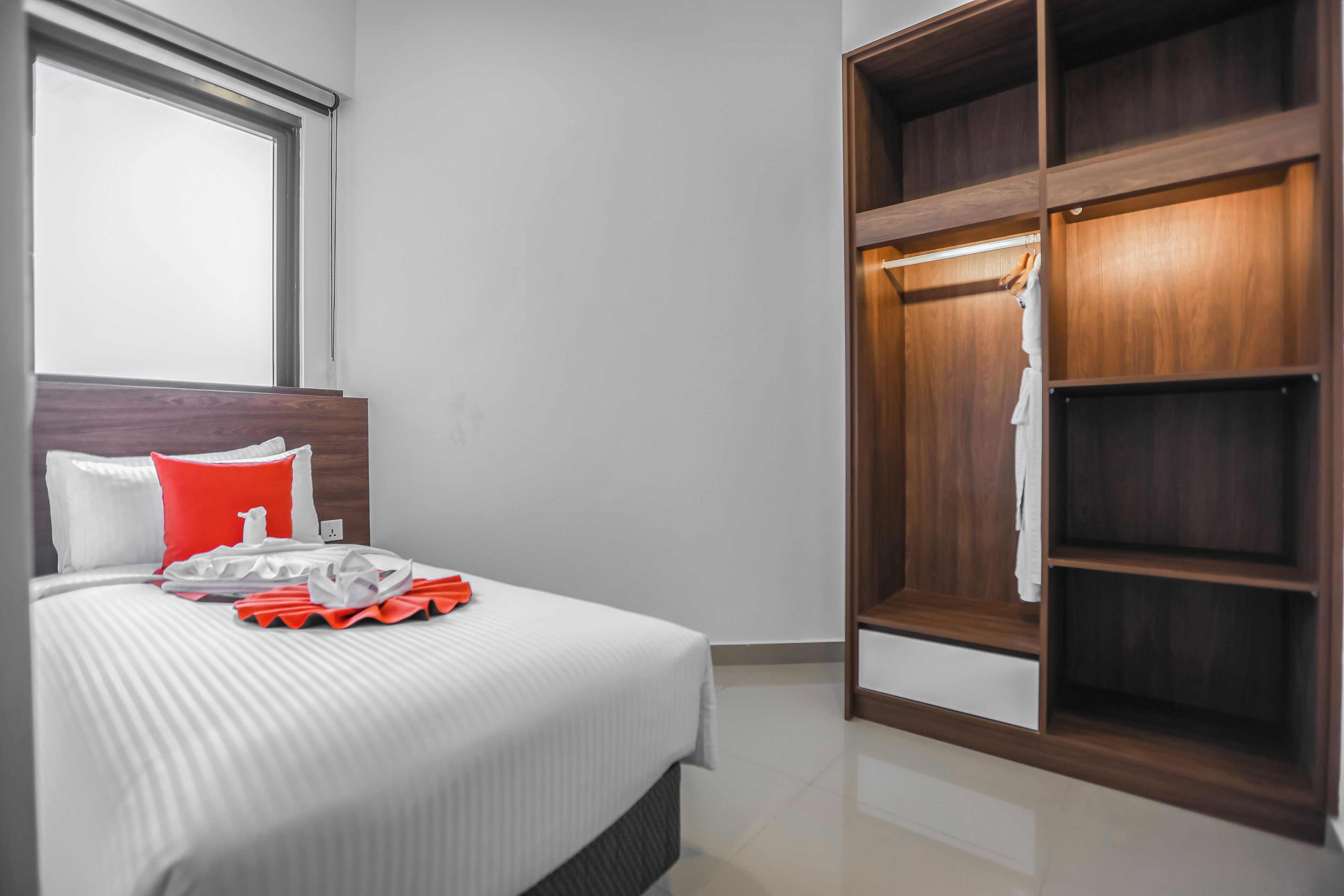 A neatly made bed with a striped bedspread, a red pillow, and a white pillow. There is a wooden wardrobe with a mirror and a hanging robe.
